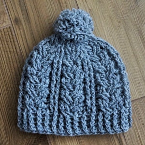 Può includere: Un berretto di lana grigio con un pompon soffice in cima. Il berretto ha un motivo a maglia testurizzato.