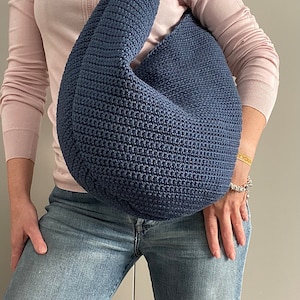 May include: A blue crocheted shoulder bag with a slouchy design. The bag is worn by a person wearing a pink sweater and blue jeans.