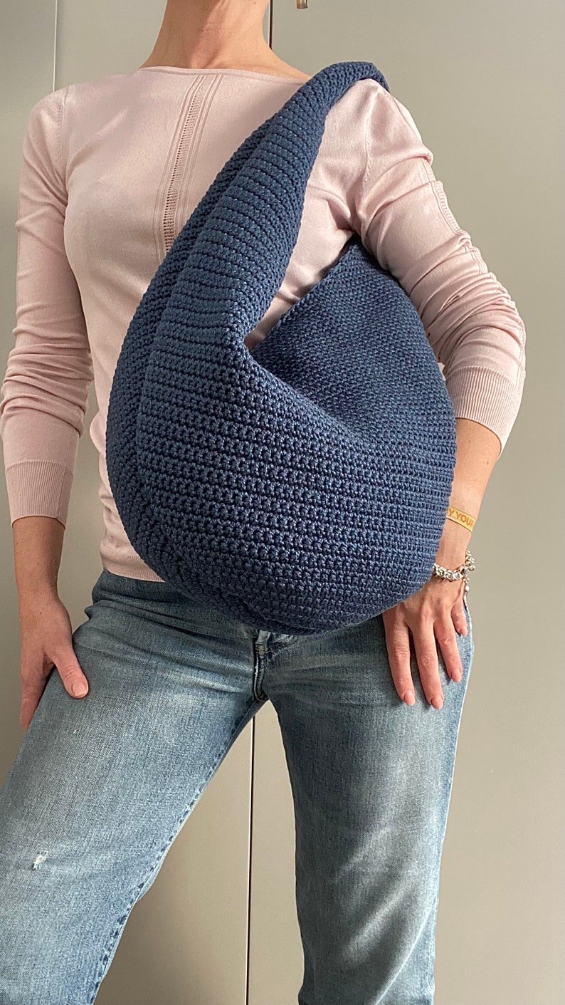 May include: A blue crocheted shoulder bag with a slouchy design. The bag is worn by a person wearing a pink sweater and blue jeans.