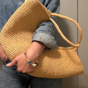 May include: A large, woven straw tote bag in a natural tan colour. The bag has a rectangular shape with a rounded top and two handles. A gold and blue beaded bracelet is visible on the wrist.