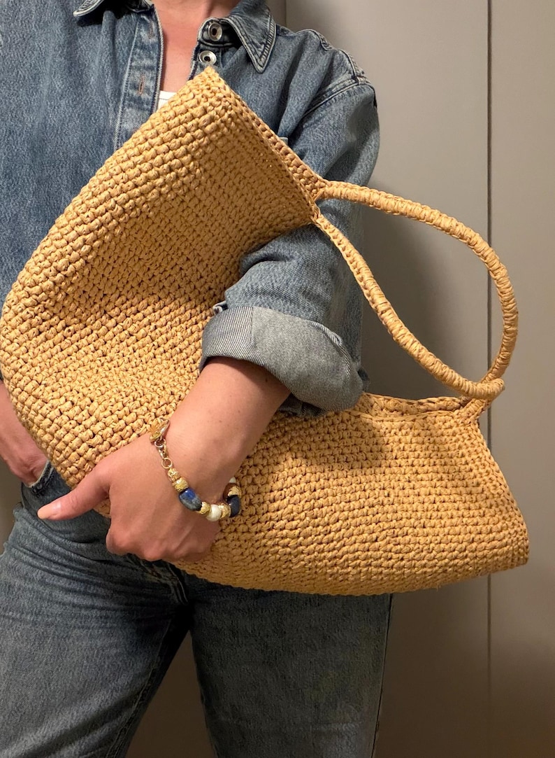 May include: A large, woven straw tote bag in a natural tan colour. The bag has a rectangular shape with a rounded top and two handles. A gold and blue beaded bracelet is visible on the wrist.