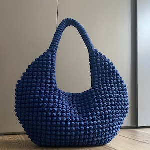 Può includere: Una borsa blu all'uncinetto con una forma arrotondata e un manico singolo. La borsa ha una superficie strutturata e irregolare, e il manico è spesso e robusto. La borsa è realizzata in un materiale morbido e flessibile.
