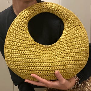 May include: Mustard yellow crochet handbag with a large circular handle. The bag is made of a textured yarn and has a rounded shape. The bag is held in front of a person wearing a black sweater.
