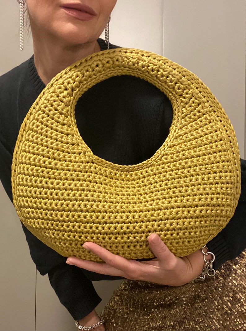 May include: Mustard yellow crochet handbag with a large circular handle. The bag is made of a textured yarn and has a rounded shape. The bag is held in front of a person wearing a black sweater.