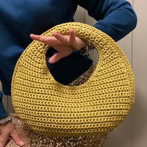 May include: A golden yellow, crocheted handbag with a large circular handle. The bag has a rounded shape and is held by a person wearing a blue sweater and a sequined skirt. The bag's texture is clearly visible, showcasing the intricate crochet work.