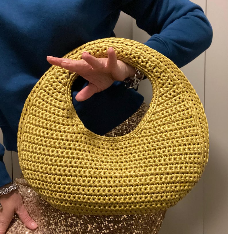 May include: A golden yellow, crocheted handbag with a large circular handle. The bag has a rounded shape and is held by a person wearing a blue sweater and a sequined skirt. The bag's texture is clearly visible, showcasing the intricate crochet work.