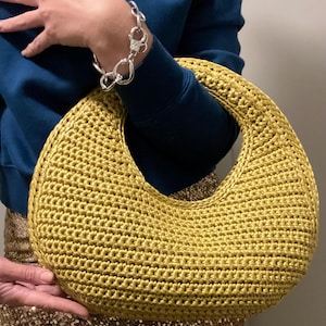Puede incluir: Un bolso de mano de ganchillo amarillo dorado en forma de media luna. El bolso tiene un asa grande y abierta y una superficie texturizada. El bolso lo sostiene una persona que lleva una sudadera azul y una pulsera plateada. El bolso es un accesorio elegante.