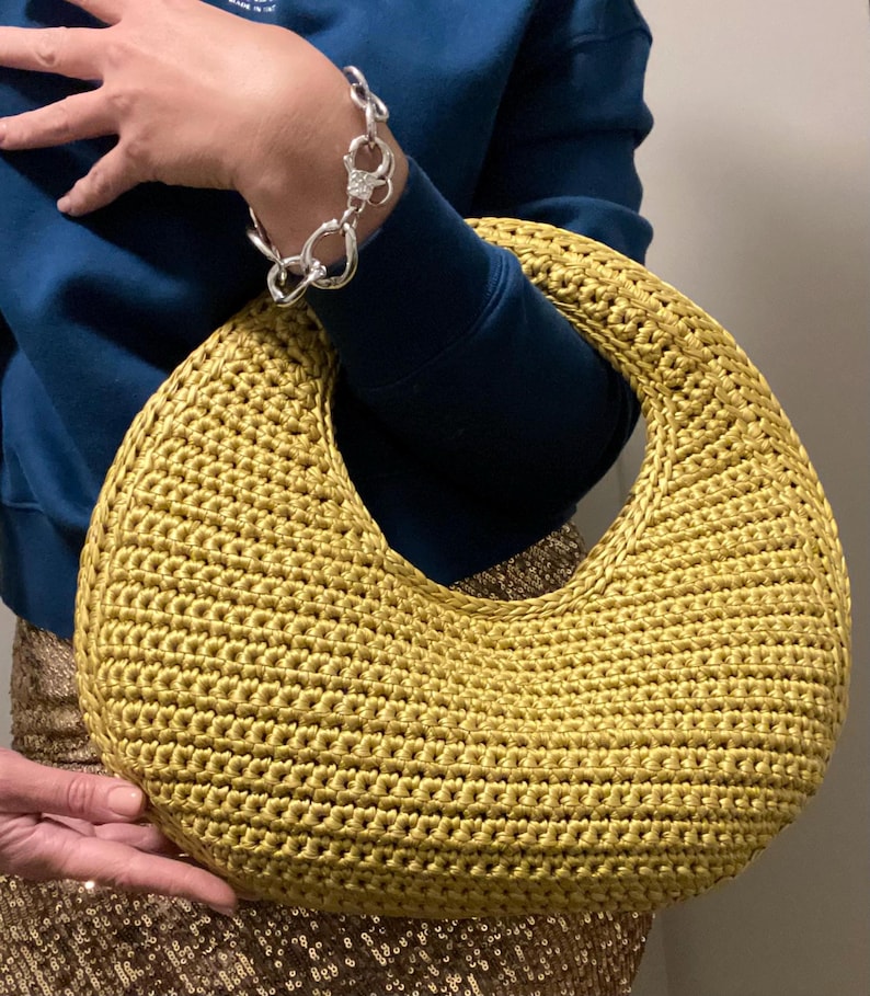 May include: A golden-yellow, crescent-shaped crochet handbag. The bag has a large, open handle and a textured surface. The bag is held by a person wearing a blue sweatshirt and a silver bracelet. The bag is a stylish accessory.