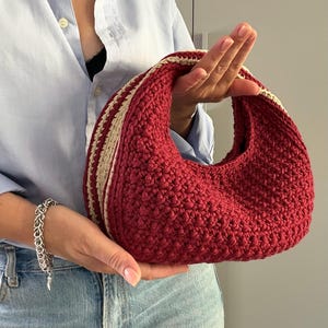 Puede incluir: Un bolso de mano en forma de media luna elaborado con hilo rojo oscuro y un patrón de punto de ganchillo texturizado. El bolso presenta una franja gruesa de color crema a lo largo del borde superior y el asa, contrastando con el cuerpo rojo.