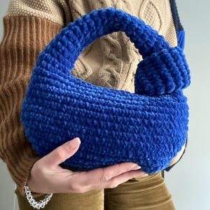 Op de afbeelding: Een blauwe gehaakte handtas met een geknoopte handvat. De tas is gemaakt van een pluche garen en heeft een zachte, textuur.