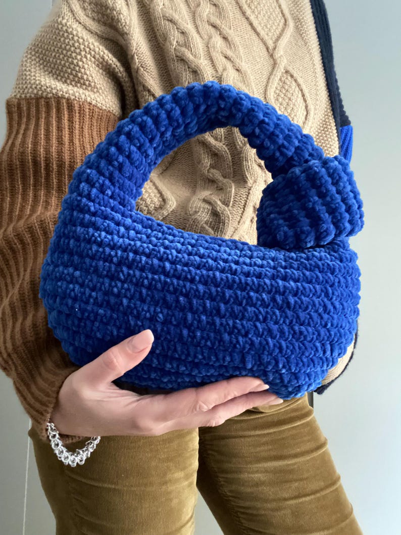 Op de afbeelding: Een blauwe gehaakte handtas met een geknoopte handvat. De tas is gemaakt van een pluche garen en heeft een zachte, textuur.