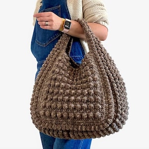 Puede incluir: Un gran bolso tote de crochet marr&oacute;n con un dise&ntilde;o texturizado y abultado. El bolso tiene una sola correa y est&aacute; hecho con un hilo grueso.