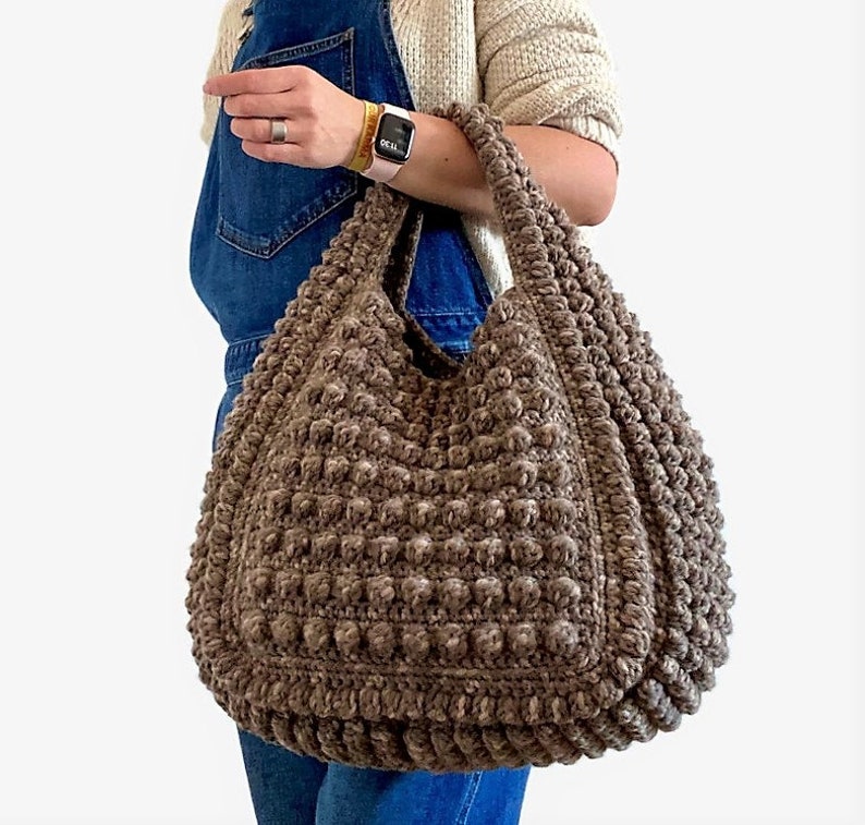 Puede incluir: Un gran bolso tote de crochet marr&oacute;n con un dise&ntilde;o texturizado y abultado. El bolso tiene una sola correa y est&aacute; hecho con un hilo grueso.