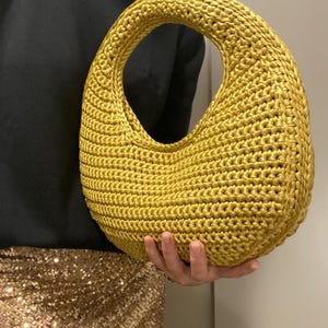 May include: A mustard yellow, crocheted handbag with a large oval opening. The bag has a rounded shape and a thick, textured handle. The bag is held in front of a person wearing a black sweater and a gold sequined skirt.