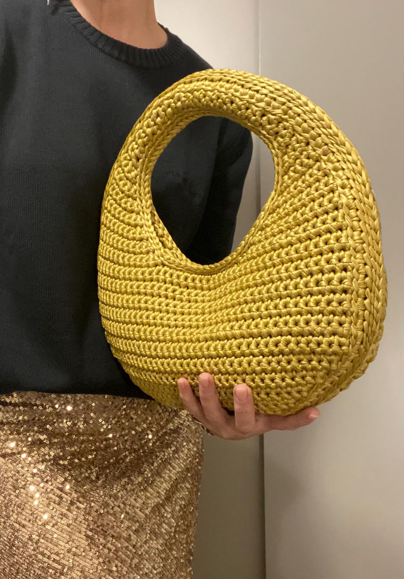May include: A mustard yellow, crocheted handbag with a large oval opening. The bag has a rounded shape and a thick, textured handle. The bag is held in front of a person wearing a black sweater and a gold sequined skirt.