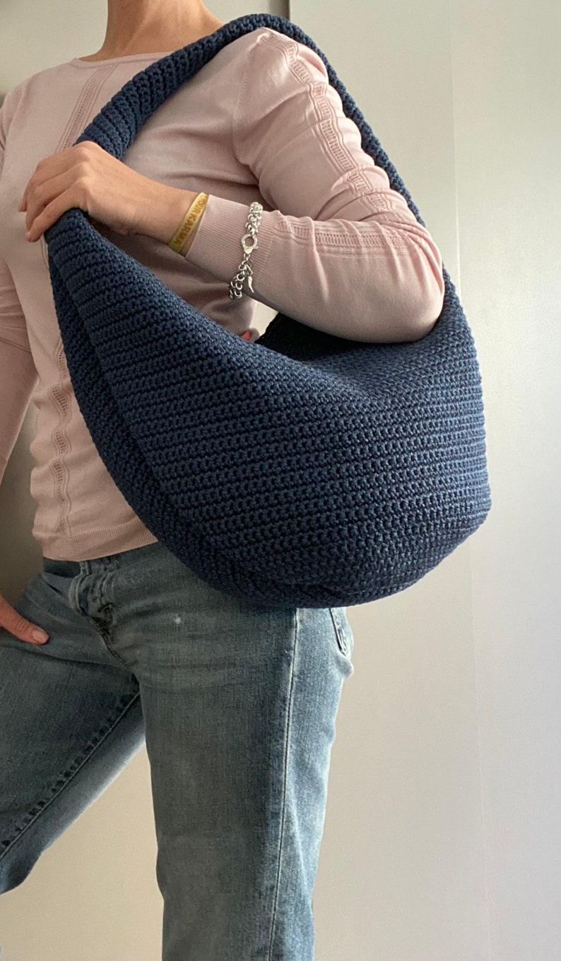 May include: A dark blue crocheted tote bag with a curved shape. The bag is being held by a person wearing a light pink sweater and blue jeans.