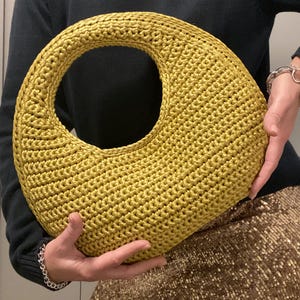 May include: A golden yellow crocheted handbag with a large circular handle. The bag has a rounded, crescent shape and is held by a person wearing a dark green sweater and a sequined skirt.