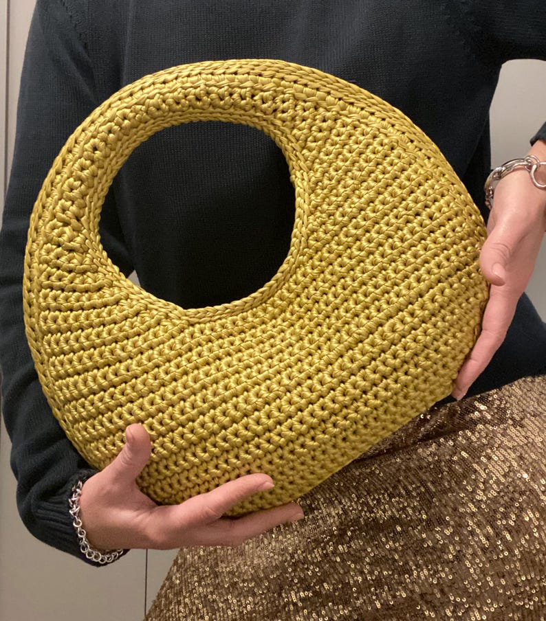 May include: A golden yellow crocheted handbag with a large circular handle. The bag has a rounded, crescent shape and is held by a person wearing a dark green sweater and a sequined skirt.