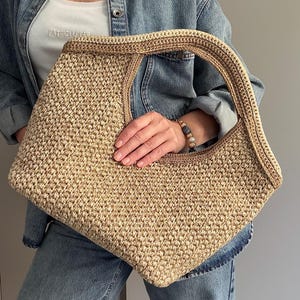 May include: A beige crocheted handbag with a large, open handle. The bag has a rectangular shape with rounded corners and a textured surface. The handle and trim are a slightly darker shade of beige.
