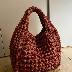 Puede incluir: Un bolso de mano de crochet marr&oacute;n con una superficie texturizada y abultada. El bolso tiene un solo asa ancha.