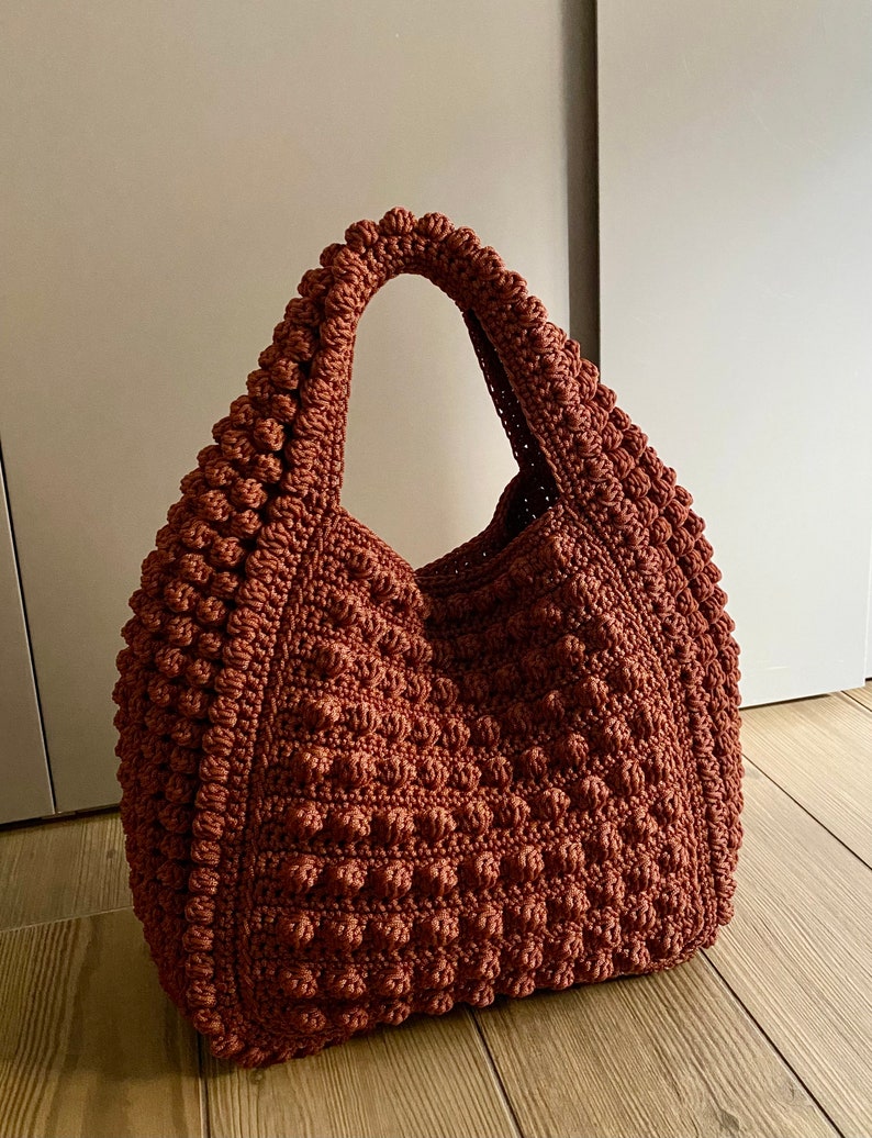 Puede incluir: Un bolso de mano de crochet marr&oacute;n con una superficie texturizada y abultada. El bolso tiene un solo asa ancha.