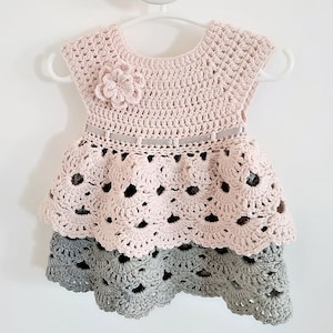 May include: A pink and gray crocheted baby dress with a flower detail and a ribbon belt.