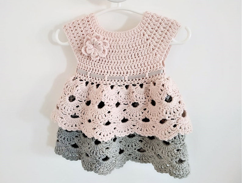 May include: A pink and gray crocheted baby dress with a flower detail and a ribbon belt.