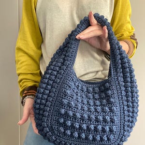 May include: A dark blue crocheted tote bag with a textured, puffy design. The bag has a single strap and is held by a person.