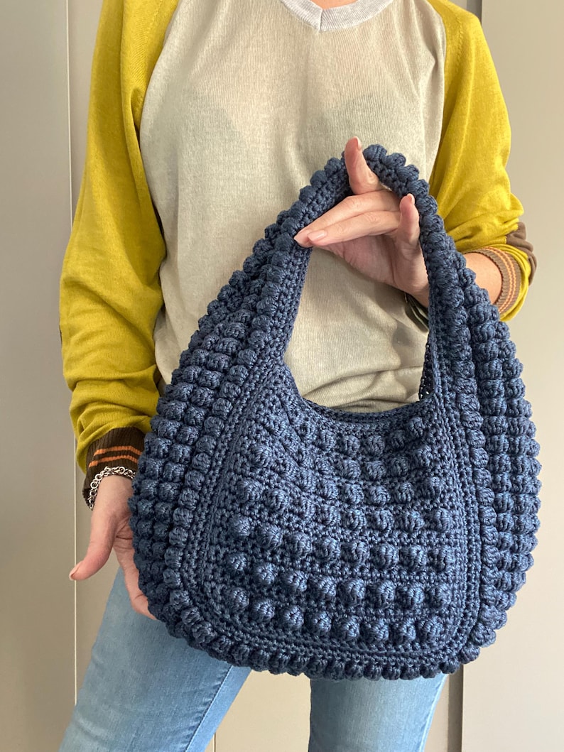 May include: A dark blue crocheted tote bag with a textured, puffy design. The bag has a single strap and is held by a person.