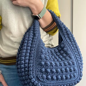 May include: A blue crocheted tote bag with a textured, bumpy design. The bag has a single strap and is worn over the shoulder.