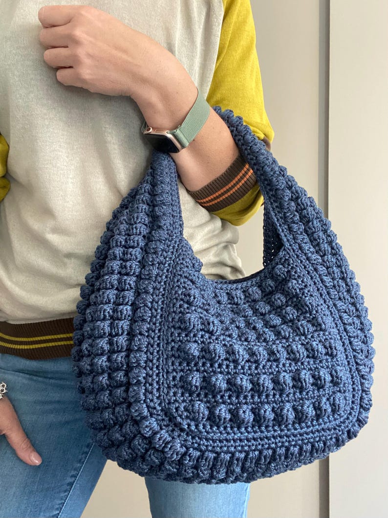 May include: A blue crocheted tote bag with a textured, bumpy design. The bag has a single strap and is worn over the shoulder.