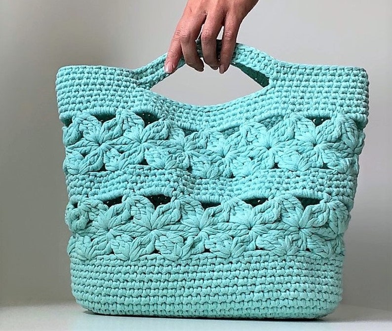 May include: A teal crocheted tote bag with a flower pattern. The bag has a wide, open top and two handles.