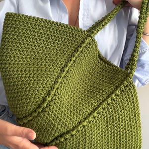 May include: A moss green crochet tote bag featuring a dense, textured stitch pattern and a prominent diagonal seam detail across the front. The bag includes two matching crocheted shoulder straps and a wide, open-top design for storage.