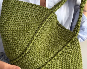 Puede incluir: Un bolso tote de ganchillo en color verde musgo con un patrón de puntos denso y texturizado, además de un detalle de costura diagonal en la parte frontal. El bolso incluye dos asas de hombro a juego y un diseño de apertura amplia.