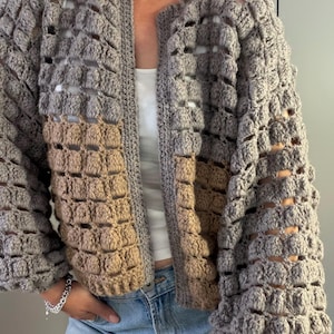 May include: A long-sleeved, open-front crochet cardigan featuring a textured bobble stitch pattern. The garment is constructed with a combination of light gray and tan yarn, showcasing a grid-like openwork design throughout the sleeves and body.