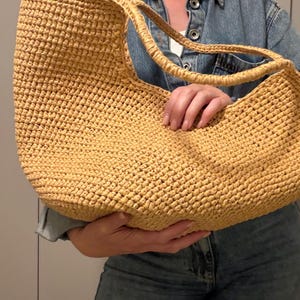 May include: A large, woven straw tote bag in a light tan colour. The bag has two handles and a rounded shape. The texture of the straw is visible, and the bag appears to be handmade. The bag is held by a person wearing a denim jacket.
