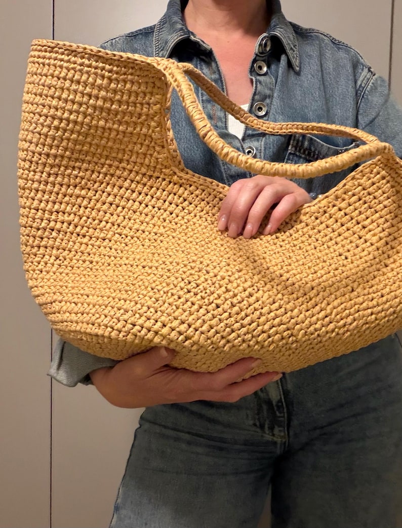 May include: A large, woven straw tote bag in a light tan colour. The bag has two handles and a rounded shape. The texture of the straw is visible, and the bag appears to be handmade. The bag is held by a person wearing a denim jacket.