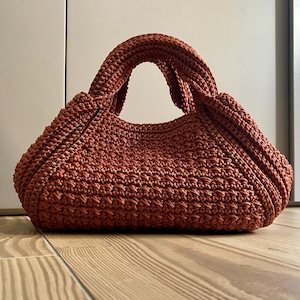 Może przedstawiać: Ręcznie robiona, rdzawa torebka z szydełka o zaokrąglonym kształcie i solidnych uchwytach. Torba ma wzorzysty ścieg. Torba jest przeznaczona do przenoszenia niezbędnych rzeczy i stoi na drewnianej podłodze.