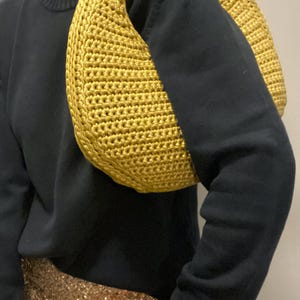 May include: A mustard yellow, crocheted shoulder bag. The bag is crescent-shaped and has a thick, rounded strap. The bag is made of a textured yarn and is worn over the shoulder. The person is wearing a black sweater and a gold sequined skirt.