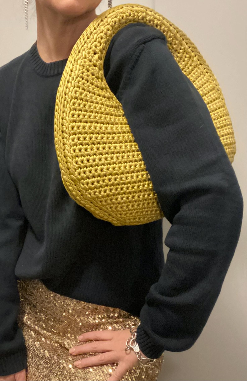 May include: A mustard yellow, crocheted shoulder bag. The bag is crescent-shaped and has a thick, rounded strap. The bag is made of a textured yarn and is worn over the shoulder. The person is wearing a black sweater and a gold sequined skirt.