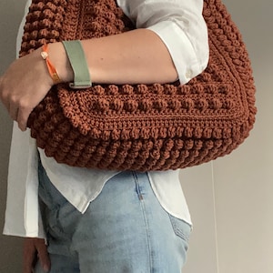 Puede incluir: Un bolso tote de crochet marr&oacute;n con una superficie texturizada y abultada. El bolso se lleva sobre el hombro de una persona que lleva una camisa blanca y unos vaqueros azules.