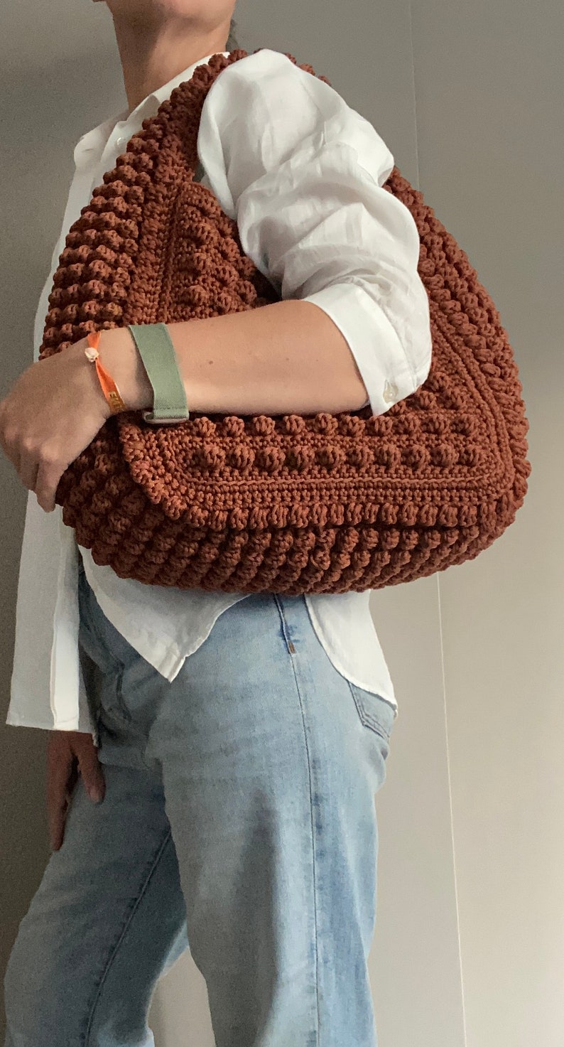 Puede incluir: Un bolso tote de crochet marr&oacute;n con una superficie texturizada y abultada. El bolso se lleva sobre el hombro de una persona que lleva una camisa blanca y unos vaqueros azules.