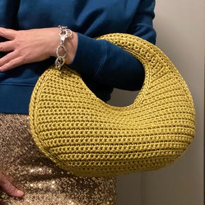 May include: A mustard-yellow, crescent-shaped crochet handbag. The bag has a rounded handle and a textured surface. A silver bracelet is visible on the arm holding the bag. The person is wearing a dark blue sweater and a gold sequined skirt.