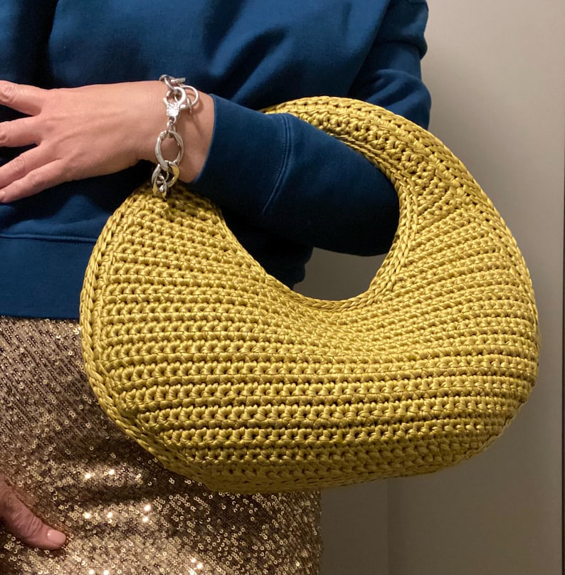 May include: A mustard-yellow, crescent-shaped crochet handbag. The bag has a rounded handle and a textured surface. A silver bracelet is visible on the arm holding the bag. The person is wearing a dark blue sweater and a gold sequined skirt.