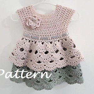 May include: A crocheted baby dress with a pink bodice and a green skirt. The dress has a flower embellishment on the bodice and a ribbon around the waist. The dress is on a hanger.