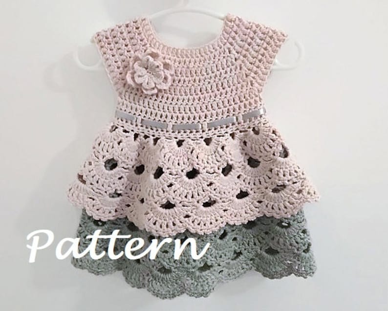 May include: A crocheted baby dress with a pink bodice and a green skirt. The dress has a flower embellishment on the bodice and a ribbon around the waist. The dress is on a hanger.