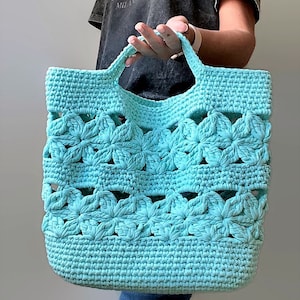 May include: A turquoise crocheted tote bag with a flower design. The bag has two handles and is made with a thick yarn.