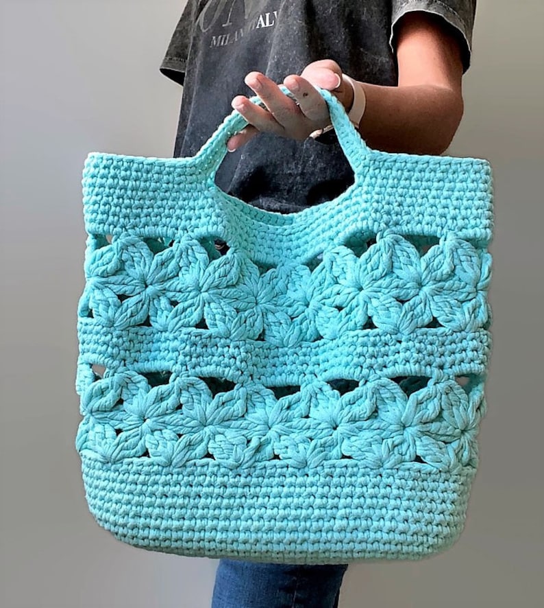 May include: A turquoise crocheted tote bag with a flower design. The bag has two handles and is made with a thick yarn.