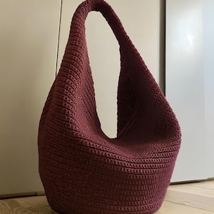 Peut inclure: Un sac en crochet bordeaux avec une seule bandoulière. Le sac a une forme arrondie et ample et est fait d'un matériau texturé et tricoté. Le sac mesure environ 46 cm de haut.