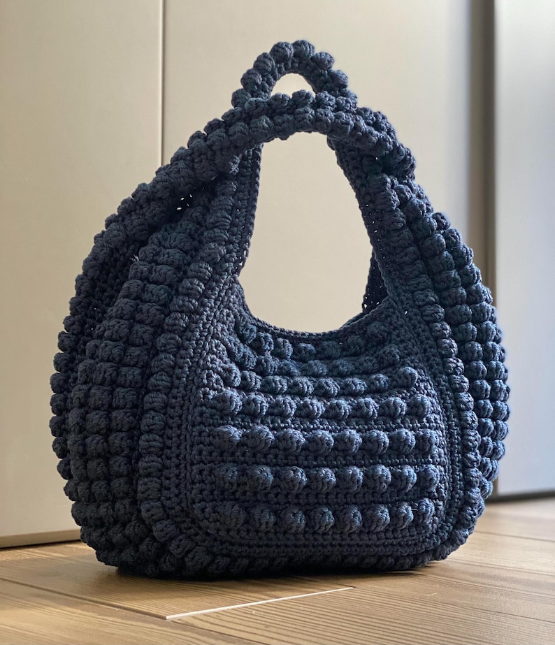 May include: A dark blue crocheted tote bag with a unique textured design. The bag has a single, large handle.